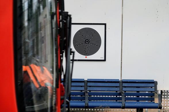 A black and white labyrinth artwork is above a bench. A tube train is passing, its flat front obscures part of the bench and one side of the frame of the labyrinth art.