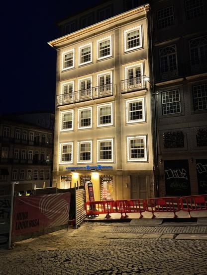 Fachadas de edifício comercial de 4 andares no Porto, à noite, com 28 janelas, cada uma circundada por 32 lâmpadas LED ligadas. O edifício destaca-se dos restantes pelo intenso brilho e poluição luminosa.