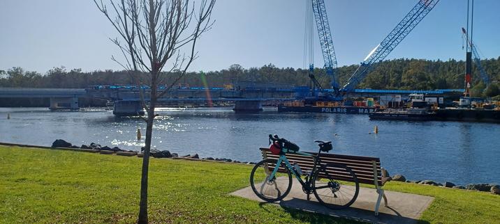 A road-bike, Minty, leans against a park bench. A large river flows under a modern bridge and cranes and other heavy equipment can be seen. A work in progress in the country town of Nelligen.