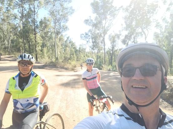The bright glare of the sun washed out a selfie of the post author and his two bike-riding friends. Large eucalyptus trees line a dirt road in the background.