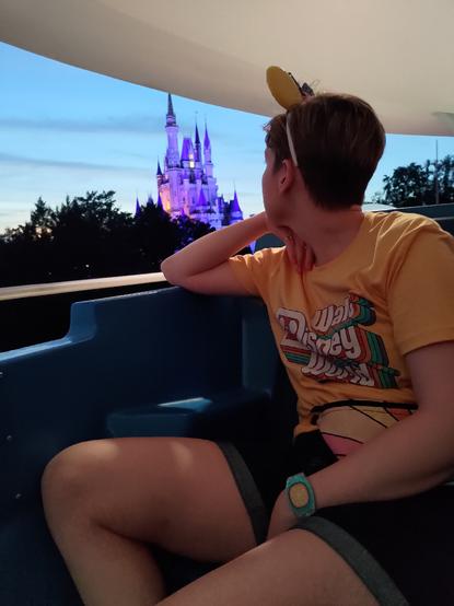 My wife looks over her shoulder towards Cinderella's castle from the Tomorrowland people mover at sunset. She is wearing a yellow Disney shirt and a pair of ears that look like the luxo junior ball.