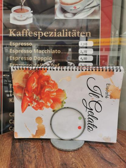 Tisch vor Eiscafe Il Gelato Eiskarte im Ständer. Dahinter das Fenster mit Aufschriften: Kaffeespezialitäten und die Preise von ESPRESSO etc.