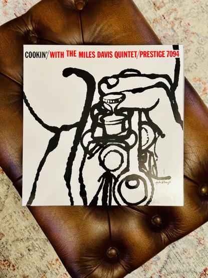 Photo of a record album jacket laying on a tan leather stool. 

The jacket is white, with a black ink sketch of fingers keying a trumpet. 

Text, in red and black, runs across the top of the jacket reading, ‘Cookin’/with the Miles Davis Quintet/Prestige 7094’.