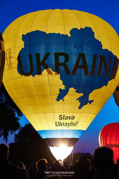 Ein gelber Heißluftballon mit den blauen, kartografischen Umrissen der Ukraine, dem Schriftzug Ukraine so wie der Botschaft "Slava Ukrayini!"
