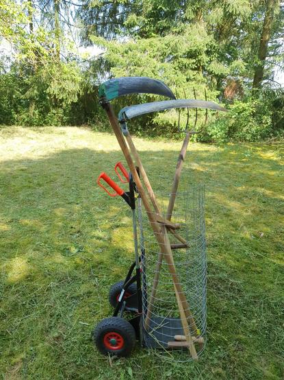Die Sackkarre steht auf der Wiese, die Estrichmatte ist mit einem Stück Draht zu einem Köcher zusammengebunden, darin stehen zwei Sensen und eine Heugabel.