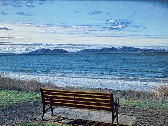 Blue rippled seas lead to the blue grey mountains. A wooden seat in the foreground has a perfect view across to the Hazards