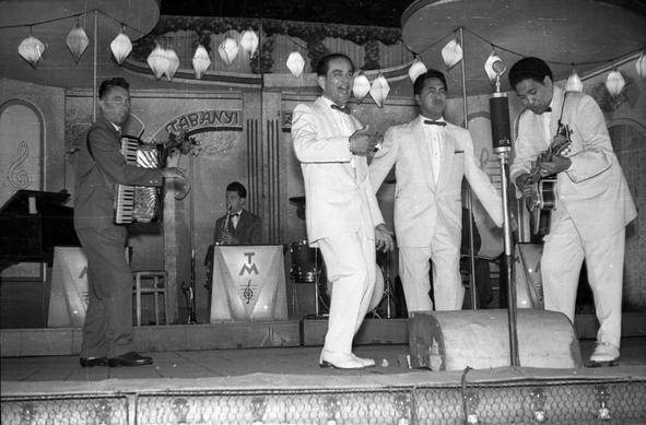 A group of musicians singing on a cabaret stage. Three are wearing white suits, one of whom is playing the guitar. There are other differently dressed musicians including an accordion player and a saxophonist also on the stage.
