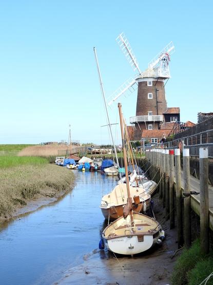 Narrow creek beside a dock with a number of small boats moored alongside. In the distance the famous mill at Cley Next The Sea.
