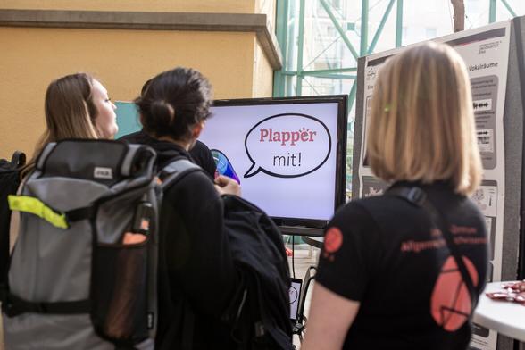 Mehrere Personen stehen vor einem Bildschirm und einem Poster. Der Bildschirm zeigt eine Sprechblase mit dem Wort "Plapper mit".