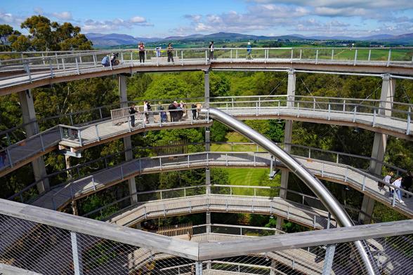 View taken at the top a viewing tower in a wooded area with mountains in the distance and some people starting a queue for an enclosed slide to the bottom of the tower.