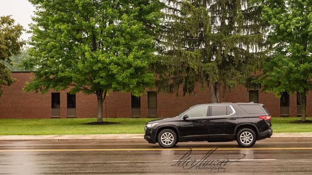 Black SUV driving on far lane.  Behind it trimmed lawn and sidewalk.  Trees and building further behind.  Cloudy sky behind the trees.