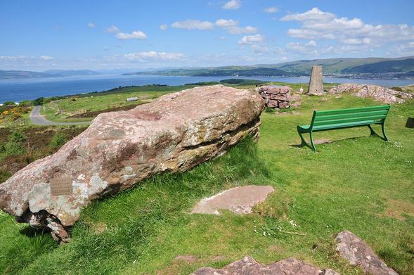 The highest point on the island of Great Cumbrae. The image shows a view taking in the distant Firth of Clyde with land visible extending across the upper part of the frame. In the foreground on the right is a trig point and a green bench, while on the left is a large whale-shaped rock in pinkish stone. The scene is in sunlight.