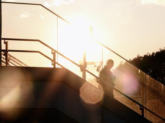 Someone walks down stairs with a glass barrier, there is bright sunlight behind them and they are just a silhouette.