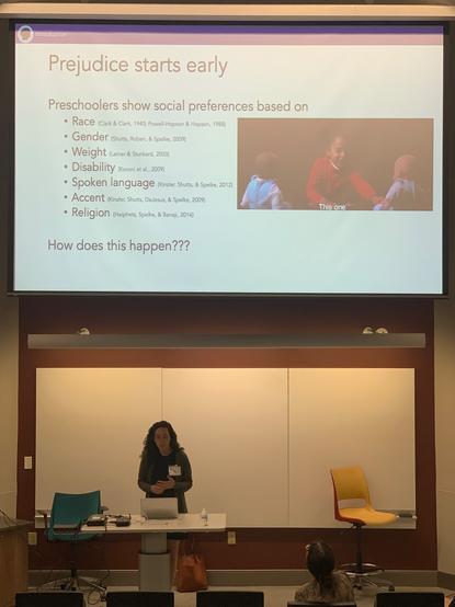 Dr. Rebecca Peretz-Lange standing below a projection of a slide listing preschoolers' documented preference/prejudice about race, gender, weight, disability, spoken language, accent, and religion.