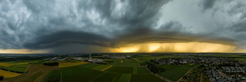 Panorama aus der Luft bei Ulm Jungingen. Am Himmel sind große, strukturierte Gewitterwolken zu sehen, der obere Bereich der Wolken und des Himmels sind grau. Der Himmel in der Mitte des Bildes ist leuchtend gelb, die untergehende Sonne scheint durch den Regen des Unwetters. Links ist im Hintergrund die Innenstadt von Ulm mit Münster zu sehen, in der Mitte das Industriegebiet 'Hörvelsinger Weg' und rechts der Stadtteil Jungingen.