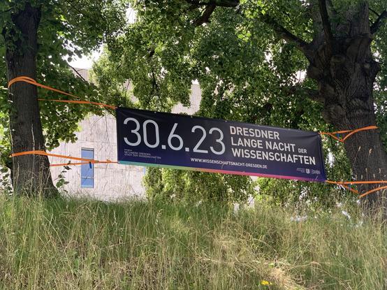 Banner mit dem Text "30.6.23 Dresdner Lange Nacht der Wissenschaften", der vor der Zentralbibliothek zwischen zwei Bäume gespannt wurde. Im Hintergrund sieht man einen Teil der Fassade der Bibliothek. Im Vordergrund ein Stück Wiese.