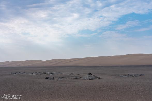 Sandy flat plain with some rocks sticking out and a row of huge dunes in the background