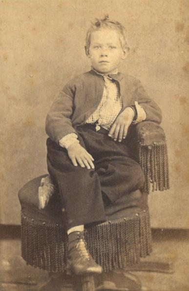 19th century photograph of a young boy lounging on a fringed stool looking very very cool. He's wearing trousers, shoes, a checked shirt and dark colored jacket.