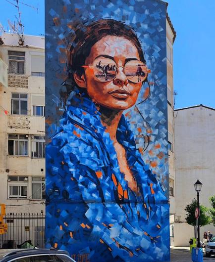 Streetartwall. On a narrow outer wall of an approximately three-story building is a wonderful portrait of a young woman with sunglasses sprayed / painted. The background consists of light and dark blue color stripes. The young dark-haired woman wears a blue blouse and is shown slightly sideways. The face, especially her mouth and narrow eyebrows are finely drawn. She wears big round sunglasses in which a scenery is reflected. She is looking at the sea. On the left you can see a fishing boat with men on the beach and on the right a man is carrying a long coiled rope to the boat. The men are setting out. The whole mural is designed in a bright blue and as if with a dabbed technique. Countless color stripes in light blue, blue and orange gives the impression of an oil painting.