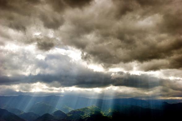 Stormy clouds are opening up, rays of light entering the cloud blanket. Green hills till the horizon.