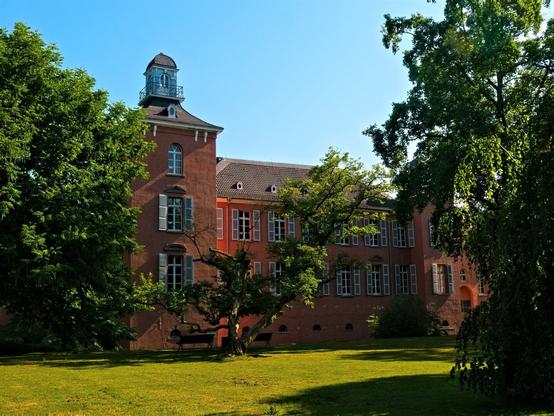 A red building with a tower surrounded by trees. In the foreground is a meadow, above it a cloudless sky.