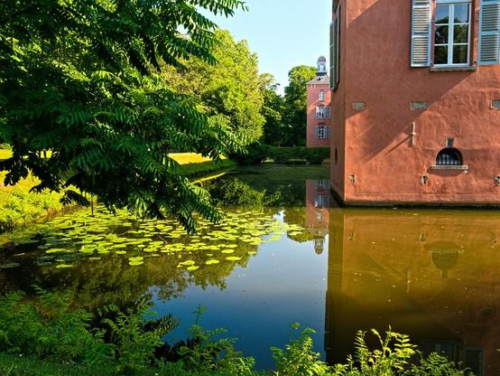 View of the moat on which the leaves of water lilies float. Parts of the red castle can be seen, including another tower. The sun shines on the moat.