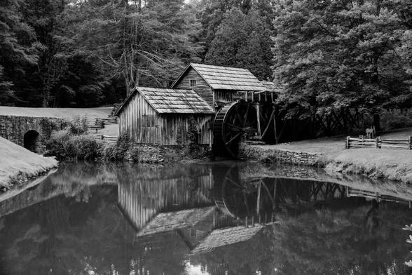 A black and white photograph of a historic wooden grist mill. The mill wheel is visible, and the mill is reflected in the pond in the foreground. Trees and grassy areas with wooden fencing surround the mill.
