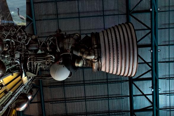 An indoor photograph, looking upward at a rocket engine. The conical exhaust nozzle is on the left side of the image, with wires and pipes leading to the connection to the gold-colored rocket bottom. There is a white sphere on the bottom of the wires and pipes. There is a corrugated metal roof in the background.
