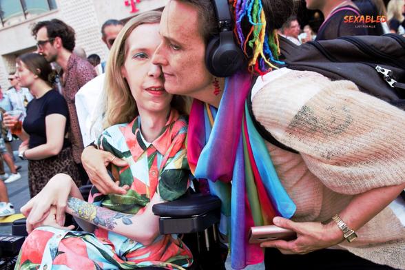 Eine Freundin umarmt mich. Ich trage ein buntes Kleid. Sie trägt einen Schal und Einsätze in Regenbogenfarben in den Haaren. Ihr Gesicht verdeckt mein müdes Lächeln zur Hälfte.