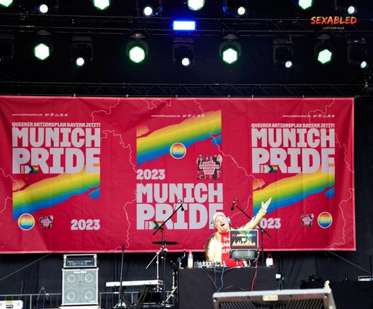DJ James Munich bei der Arbeit auf der Hauptbühne. Im Hintergrund befindet sich das rötliche Banner des CSD mit dem Motto "Queerer Aktionsplan Bayern jetzt!"