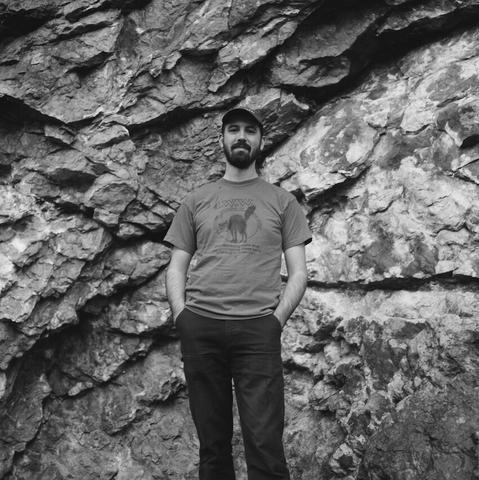 A black-and-white photo of composer and musician Andrew Weathers standing in front of some distinctive rocks. Could be the red rocks in Colorado or New Mexico. He’s a tall man with a beard wearing a baseball cap, t-shirt, and dark work pants.