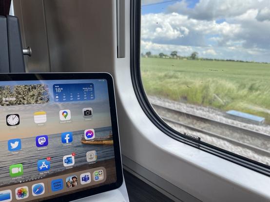 View through a train window to green fields. To the left of the picture is an iPad screen on a desk.