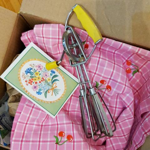Offener Pappkarton, darin liegt eine rosa-karierte Tischdecke mit aufgestickten Kirschen. Darauf links eine wundervoll altmodische Karte mit Blümchen und rechts ein metallener Handmixer mit senfgelbem Handgriff.