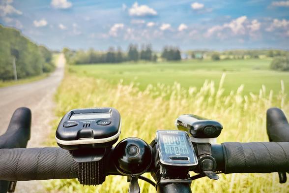The bars of a gravel bike with a farm country valley in the distance and a gravel road pointing off to the horizon on the left. It’s a hot muggy day and in the photo it looks it. In the foreground, the tall amber grasses typical of many roadsides here.
