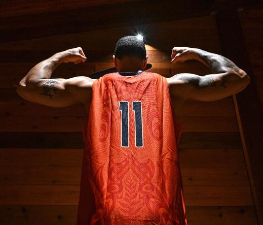 A basketball player's back is toward the camera. He is flexing his arms on each side. He is wearing an orange jersey with the designs of cedar branches and Coast Salish styled Foxes touching noses at the centre back on the jersey.