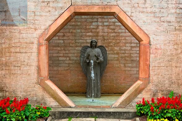 An outdoor, daylight photograph of an octagonal opening in a brick wall, revealing an angel statue standing over a fountain in a pool of water. Red flowers are at the bottom of the image except for the stair that leads to the fountain.