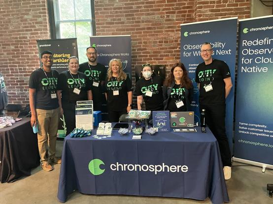 Group of Chronosphere employees, including yours truly, in matching O11Y Legend t-shirts, standing behind our nicely setup booth filled with awesome swag.