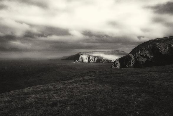 A black and white image of the headland at Cape Wrath in Scotland. It is winter and the light is fading. In the distance the headland is illuminated by sunlight