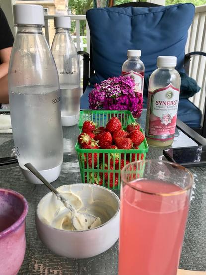 A glass patio table with bottles of still and sparkling water, kombucha, green plastic punnet box filled with heliotrope and phlox purple flowers there's a phlox purple and white bouquet of pinks (i think) in there two. There' s also a punnet o fresh strawberries, and  bowl of thickened cream sweetened with brown sugar with a dash of salt. In the foreground are a pink ceramic cup and a glass with rhubarb cordial.
