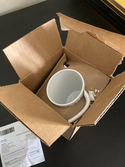 Photograph of a mug still in cardboard delivery packaging with a shattered handle.