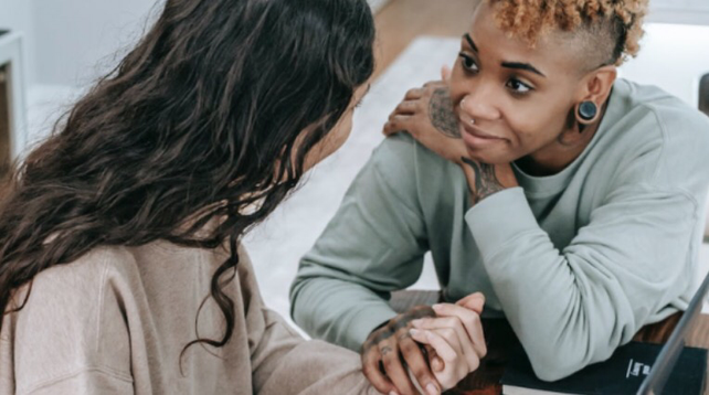 We zien een kleurenfoto. Hierop zitten twee personen. Ze houden elkaars hand vast. Een is een persoon van kleur en van de ander is dat niet zo goed te duiden. De persoon van kleur kijkt de ander in de ogen met een mooie, open blik. Die draagt een long sleeves sweater, heeft diens nek en handen gecoverd met tattoos, de oorlel gestretcht en een zwarte piercing, kort krullend lichtbruin afro haar. De andere persoon zien we deels op de rug. Die heeft lang zwart licht golvend haar.