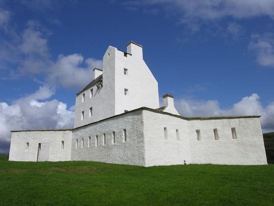 Corgarff Castle. The image shows a grassy foreground leading to a white-painted star-shaped wall pierced by rifle slits. Contained within the wall is a white-painted tower house that rises above the wall. The castle is in sunlight.