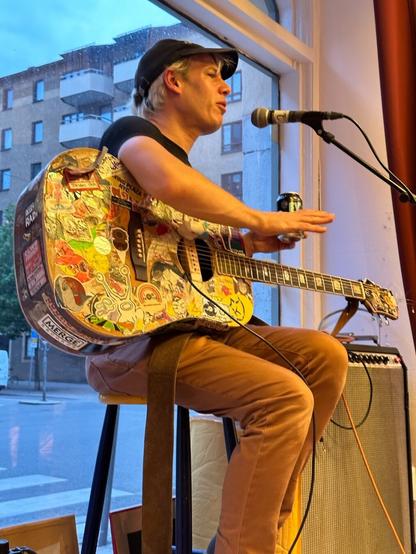 Jeffrey Lewis, a white man in brown jeans, a black T-shirt and black baseball cap, sitting on a stool in a shop window, sticker-covered acoustic guitar on his lap, holding a can of beer. Behind him, outside the window, is a streetscape of Swedish apartment buildings.