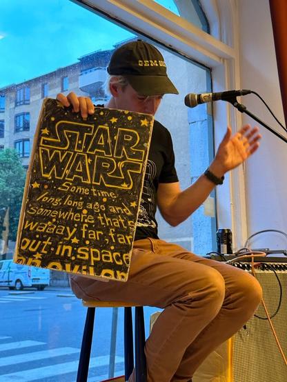 Jeffrey Lewis, a white man in brown jeans, a black T-shirt and black baseball cap, sitting on a stool in a shop window, showing a pad of paper with a hand-drawn replica of the Star Wars opening credits on it.