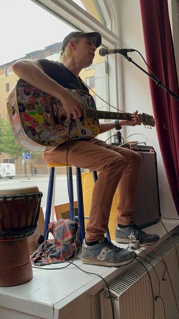Jeffrey Lewis, a white man in brown jeans, a black T-shirt and black baseball cap, sitting on a stool in a shop window, playing an acoustic guitar and singing into a microphone. A bongo drum stands next to his chair.