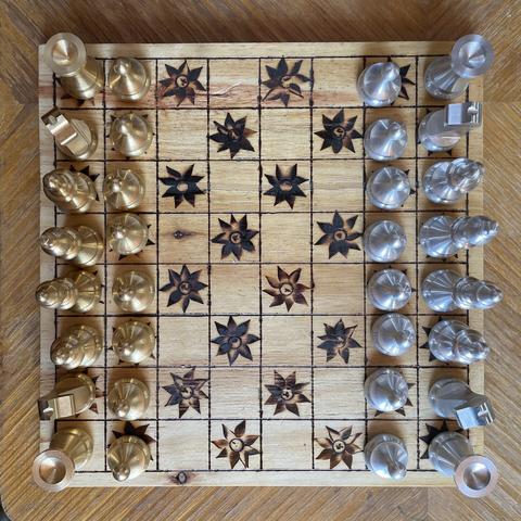 Overhead view of a chess board with a silver and brass colored sets. The black squares on the chess board are denoted by irregular star shapes burnt into the chess board.