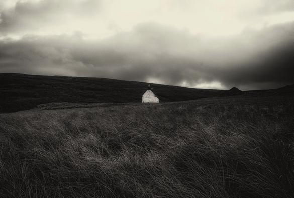 A lone building sits nestled against the hills. The image is in black and white with the slightest of sepia tinges. The mood is of isolation and desolation
