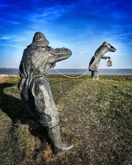 Two male statues stand on the dyk and look out to the North Sea, one with a lamp in his hand, the other securing the first one with a rope.