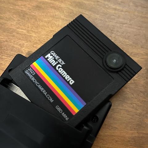 A Game Boy Mini Camera - a Game Boy Camera redesigned into a typical Game Boy cartridge. It’s hanging out the back of a black Game Boy Pocket
