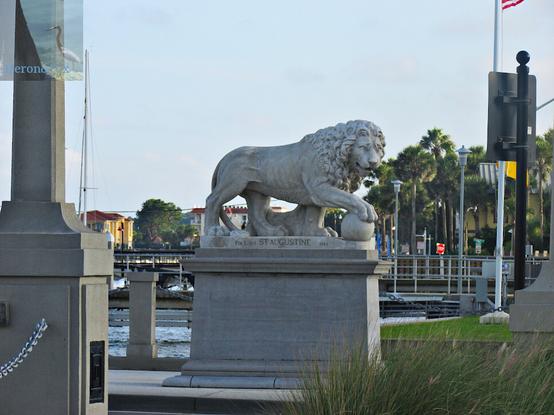 An outdoor photograph of a marble male lion on a tall marble pedestal. The statue is on a city street.